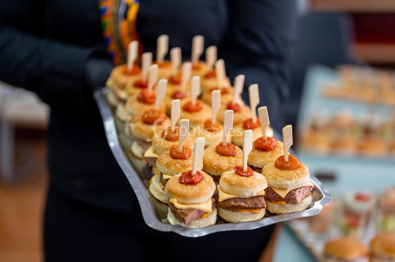 Plateau de mini-burgers garnis de sauce et maintenus par des piques en bois, tenu par une personne en tenue noire.