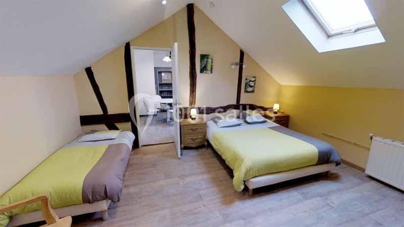 Chambre mansardée avec deux lits, parquet, poutres apparentes, éclairage doux et une porte ouverte sur une autre pièce.