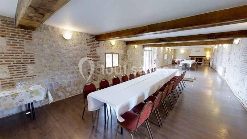 Salle en pierre avec poutres apparentes, grande table rectangulaire dressée avec nappes blanches et chaises rouges.