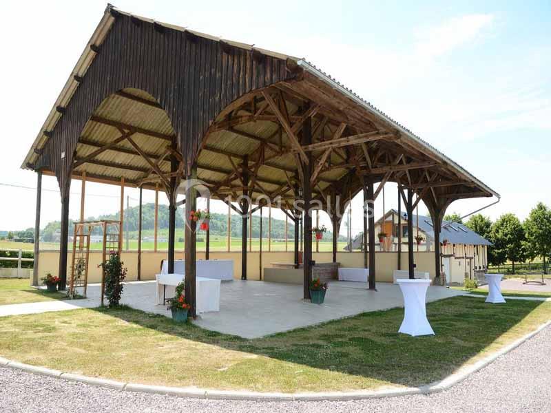 Structure en bois ouverte avec toit en tôle, entourée de verdure, utilisée pour des événements en extérieur.