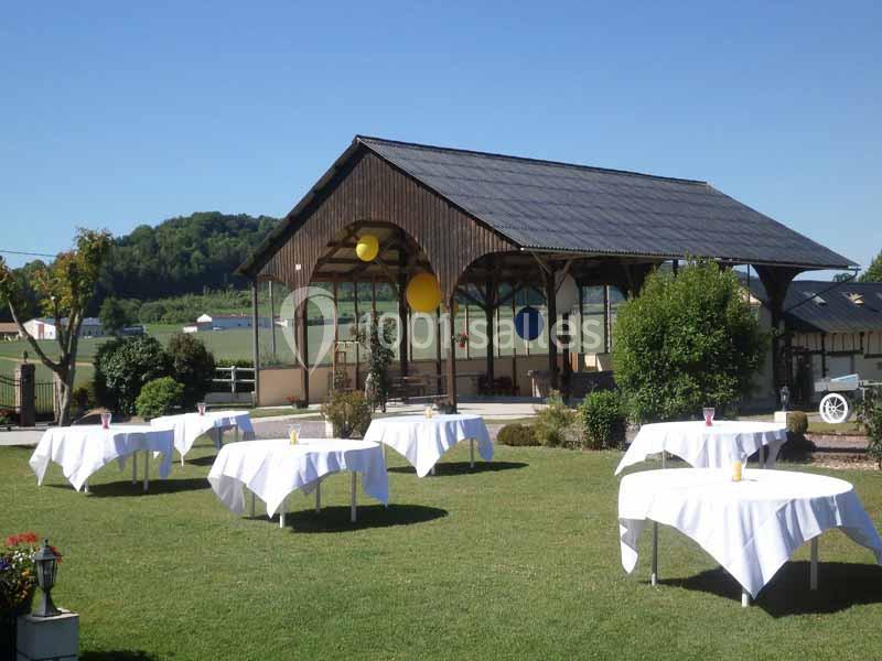 Tables rondes dressées avec nappes blanches sur une pelouse, devant une structure en bois ouverte dans un cadre champêtre.