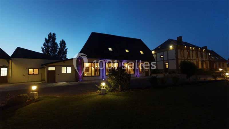 Bâtiment éclairé avec des lumières extérieures, vu de nuit, entouré d'un jardin et d'un ciel sombre.