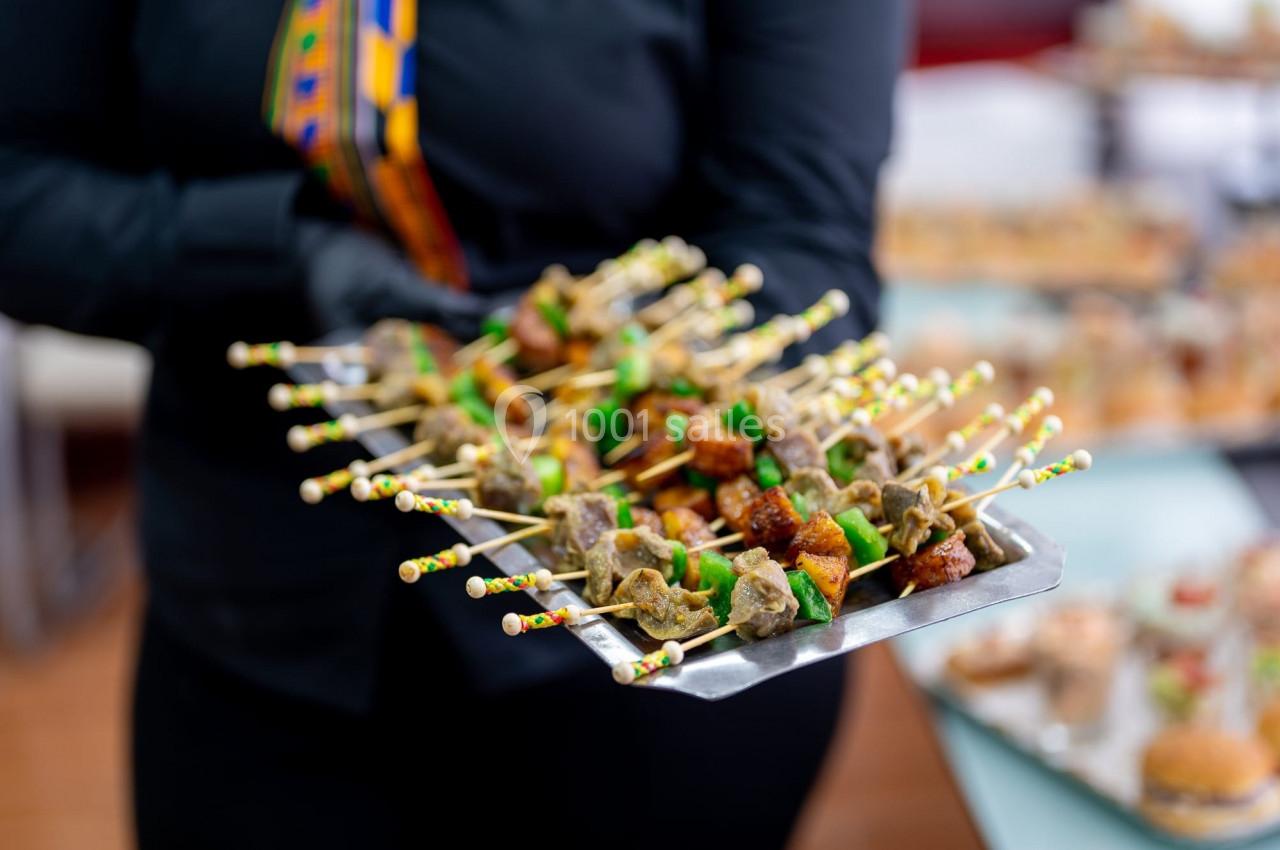Plateau de brochettes apéritives colorées, tenues par une personne vêtue de noir avec un détail de tissu coloré.