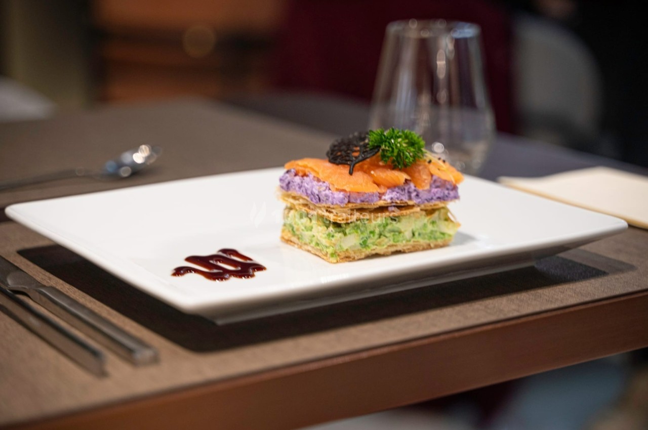 Millefeuille salé avec couches de légumes et saumon, décoré de caviar et d'herbes, servi sur une assiette blanche.