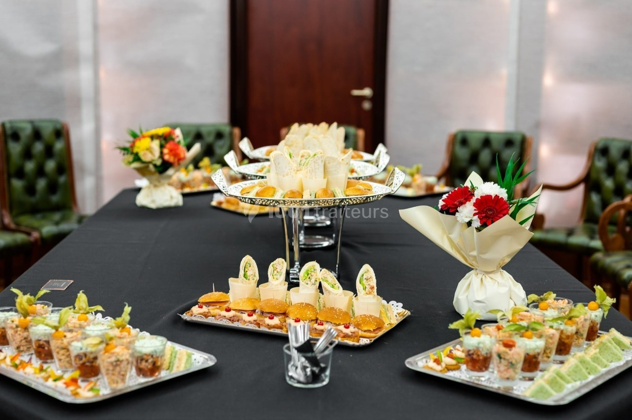 Table garnie de plateaux de bouchées apéritives variées, entourée de chaises et décorée de bouquets de fleurs.
