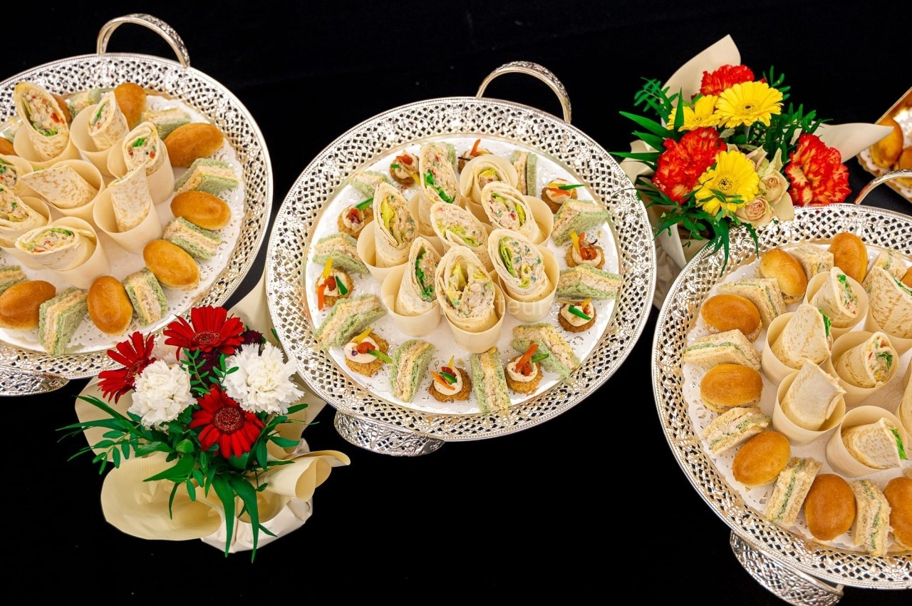 Plateaux garnis de sandwichs variés, roulés et amuse-bouches, entourés de bouquets de fleurs colorées.