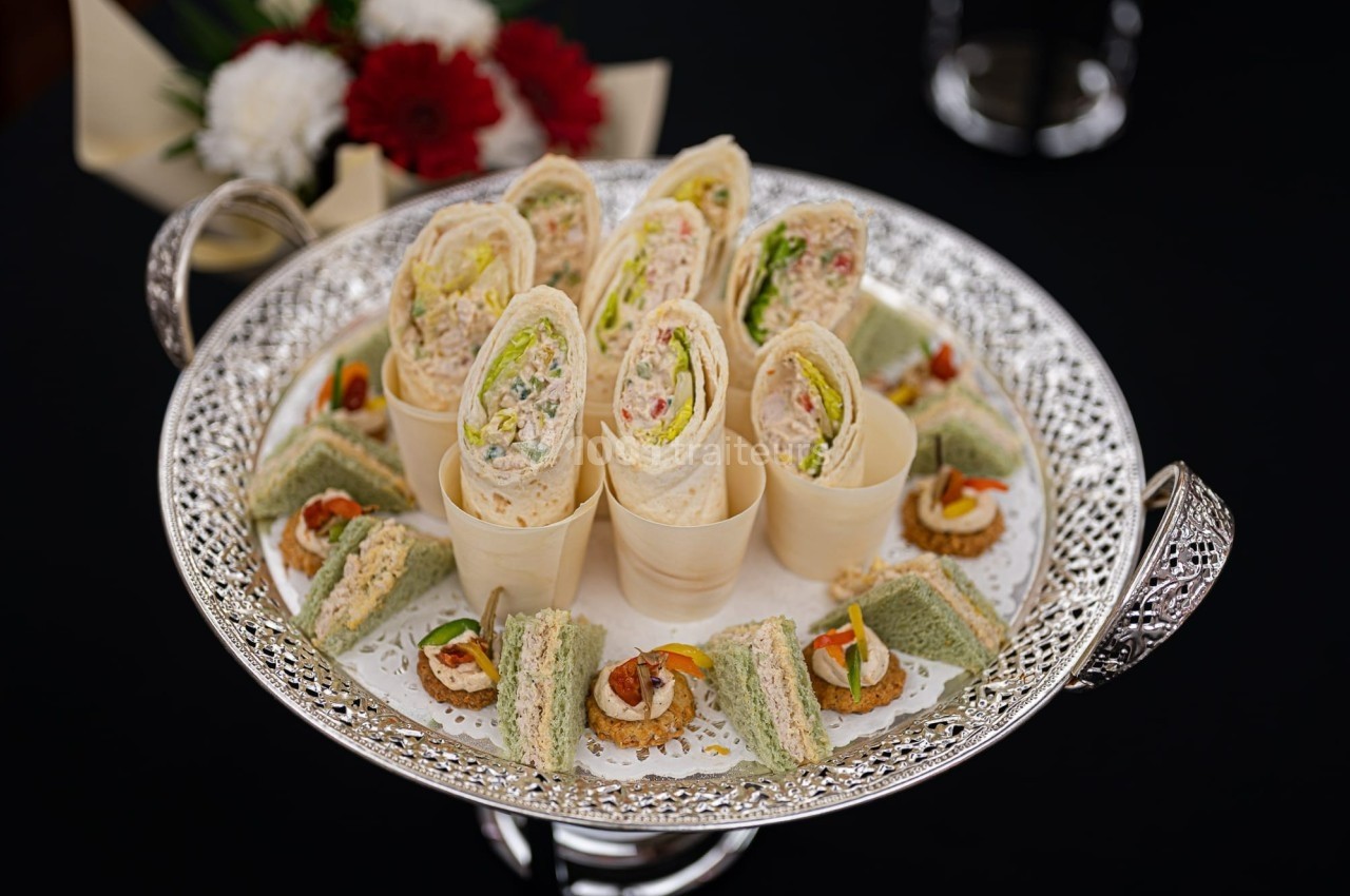 Plateau argenté avec des roulés de tortillas, des sandwichs triangulaires et des canapés sur une nappe noire.