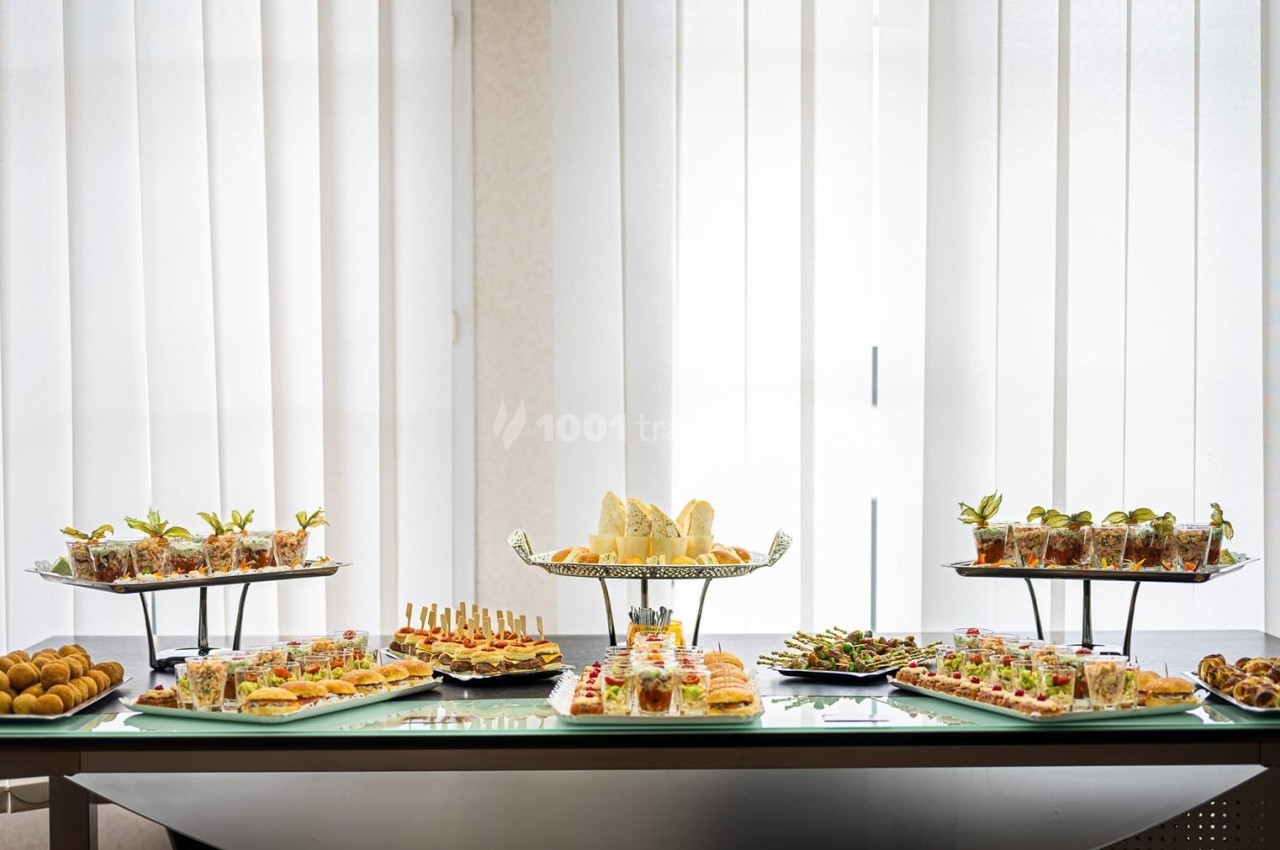 Buffet de table avec divers plats et amuse-bouches présentés sur des plateaux devant une grande fenêtre avec rideaux.