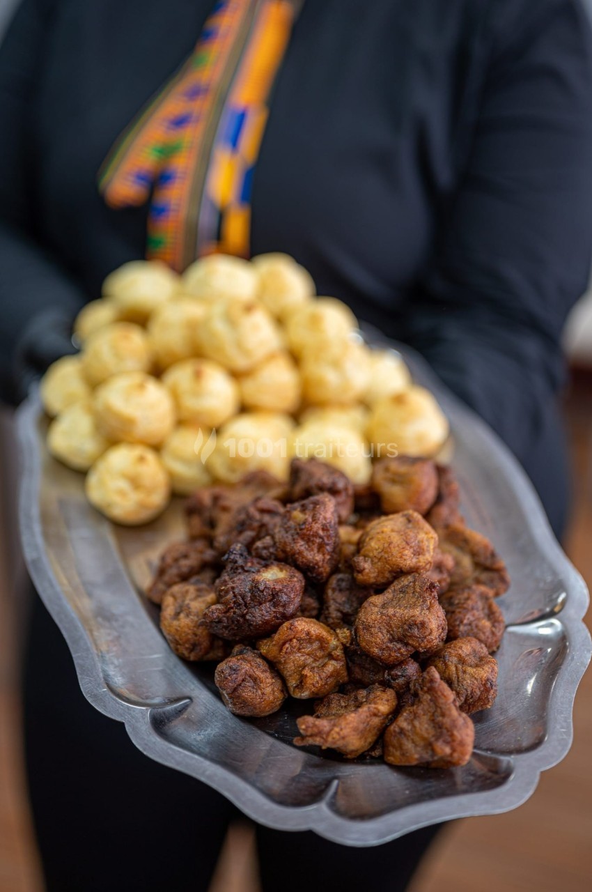 Plateau en métal contenant des beignets dorés et des boulettes frites, tenu par une personne en tenue sombre.