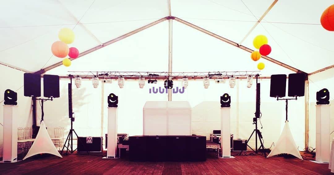 Scène de DJ sous une tente blanche, équipée de haut-parleurs, lumières et décorations suspendues colorées.