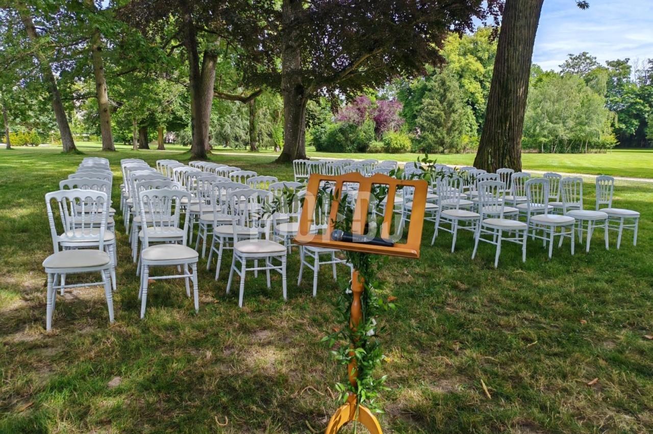 Chaises blanches alignées en extérieur sous des arbres, avec un pupitre en bois décoré de feuillage au premier plan.