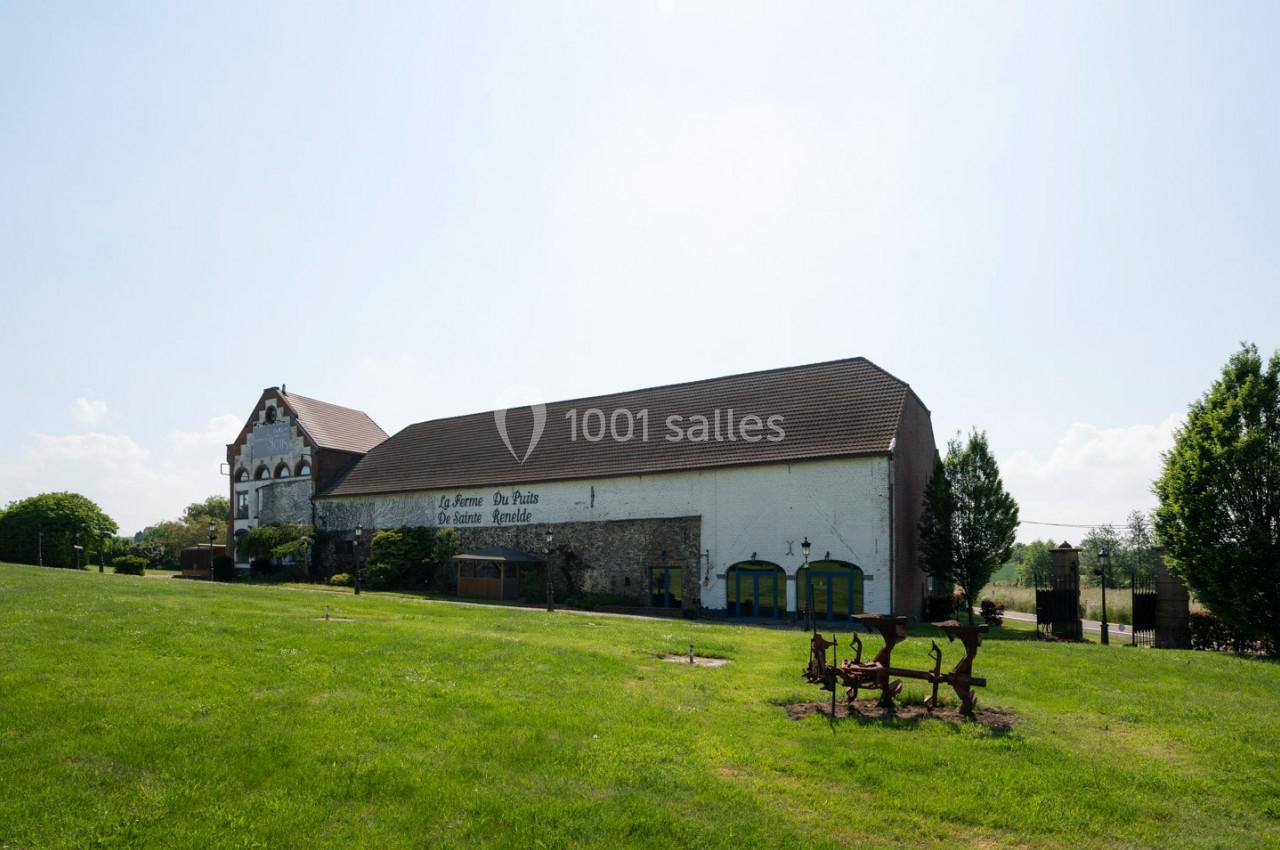 Bâtiment ancien avec toit en pente, entouré de pelouse et d'arbres, sous un ciel clair et ensoleillé.