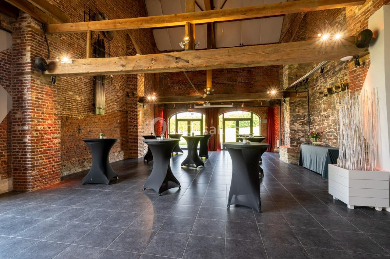 Salle en briques avec poutres apparentes, tables hautes noires et grandes ouvertures donnant sur un espace extérieur.