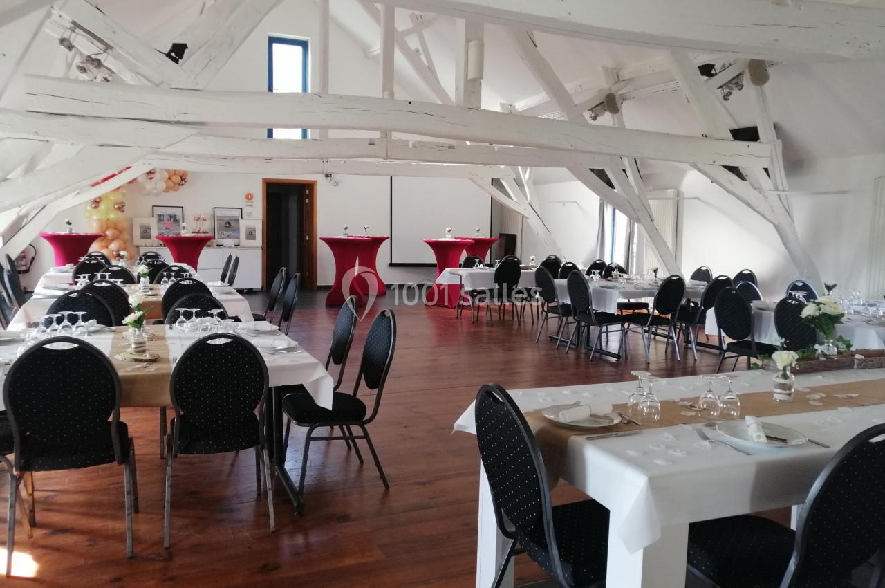 Salle lumineuse avec poutres apparentes, tables dressées pour un événement et chaises noires disposées autour.