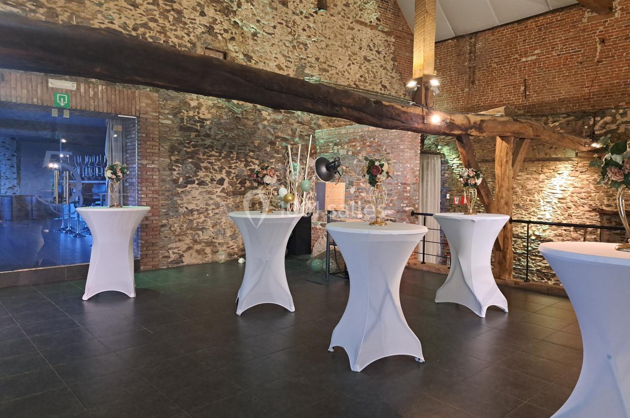Salle en pierre avec des tables hautes drapées de nappes blanches, décorées de fleurs, éclairage chaleureux.