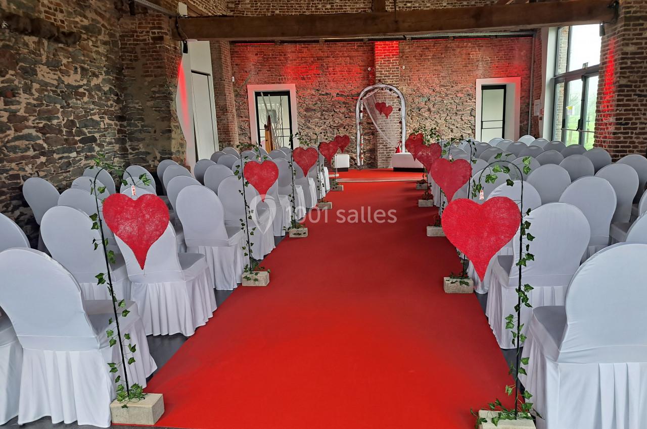 Salle de cérémonie décorée avec des chaises blanches, un tapis rouge et des cœurs rouges sur des arches végétales.