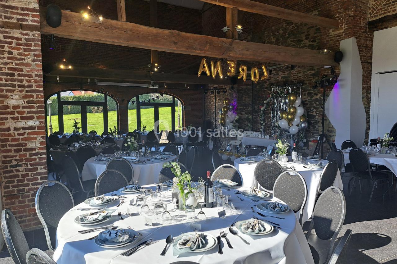 Salle de réception décorée avec des tables rondes dressées, des ballons et une vue sur un jardin à travers de grandes baies…