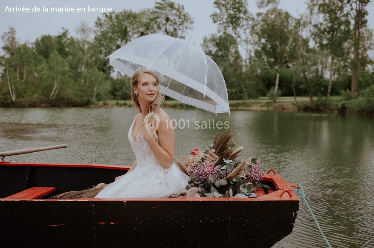 Une mariée en robe blanche, assise dans une barque sur un étang, tenant un parapluie transparent.