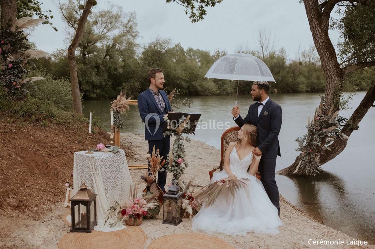 Un couple assis sous un parapluie lors d'une cérémonie laïque en plein air près d'une rivière.
