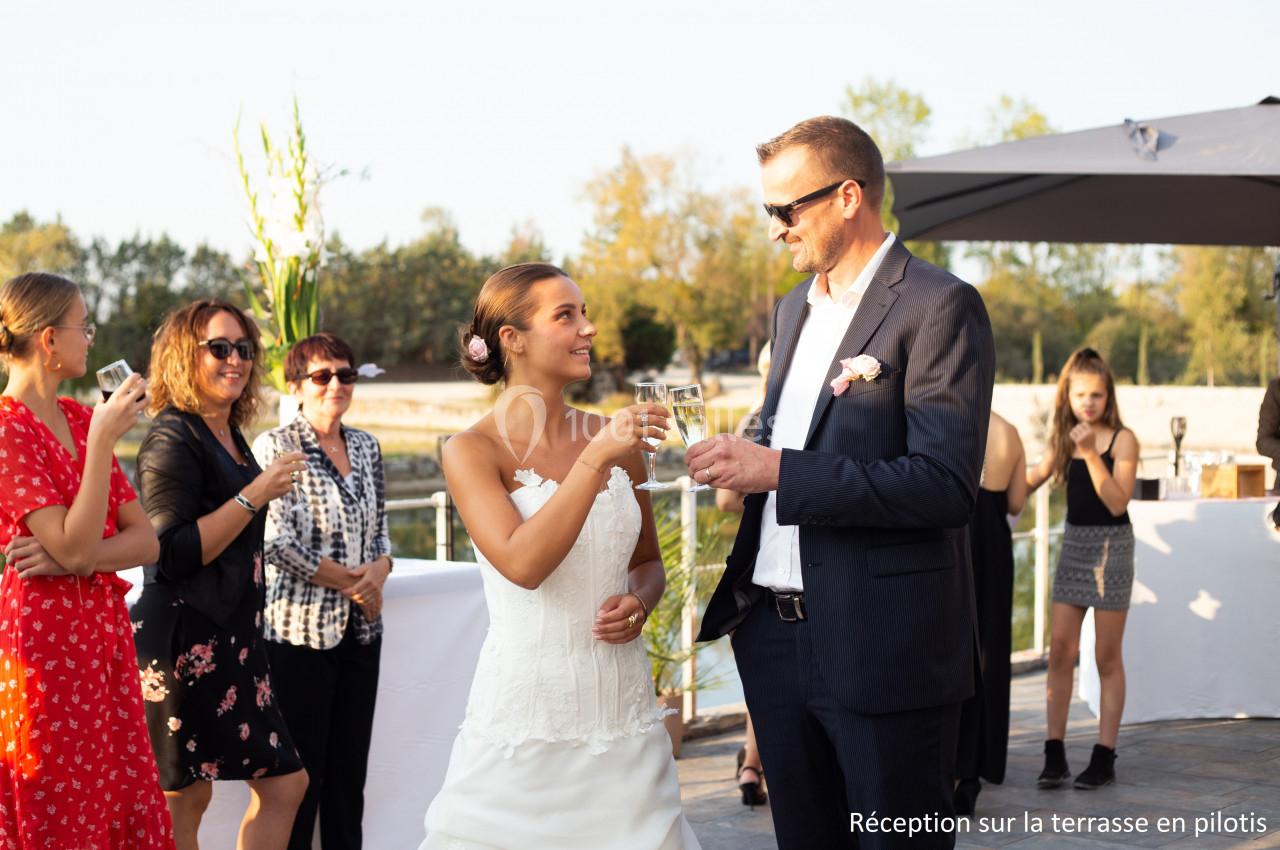 Une mariée et un homme en costume trinquent lors d'une réception en extérieur sur une terrasse ensoleillée.