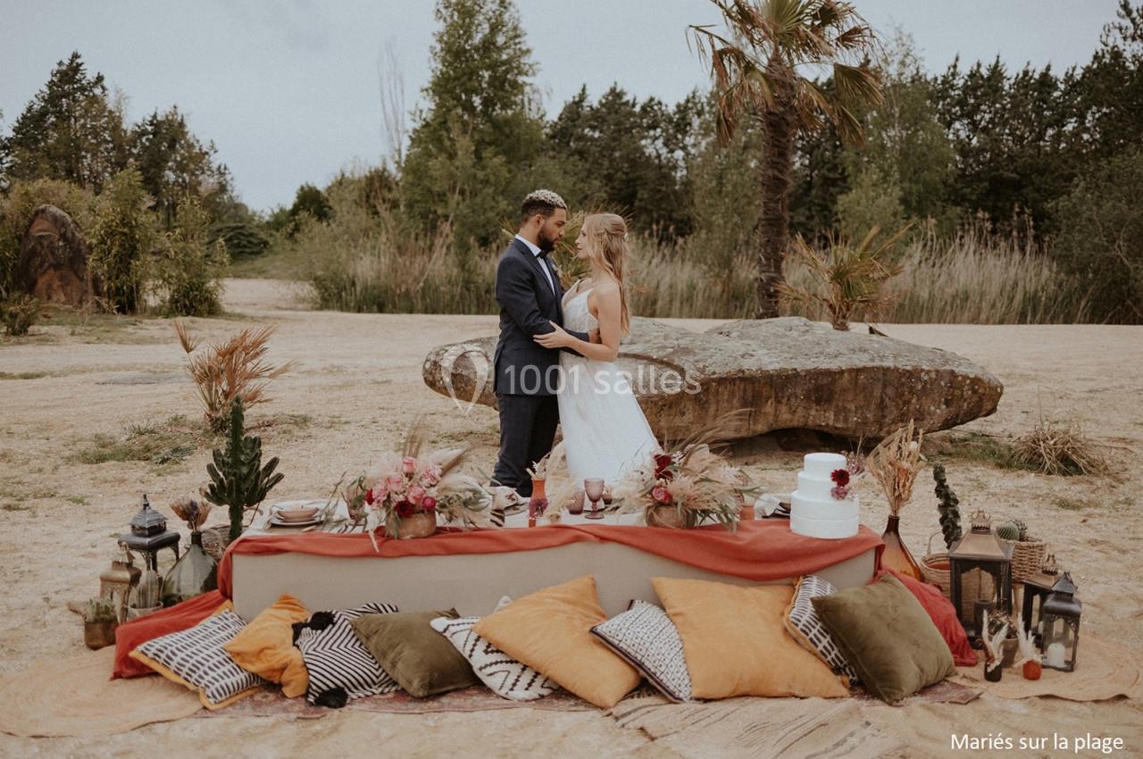Un couple en tenue de mariage se tient devant une table décorée sur une plage avec des coussins et des plantes.