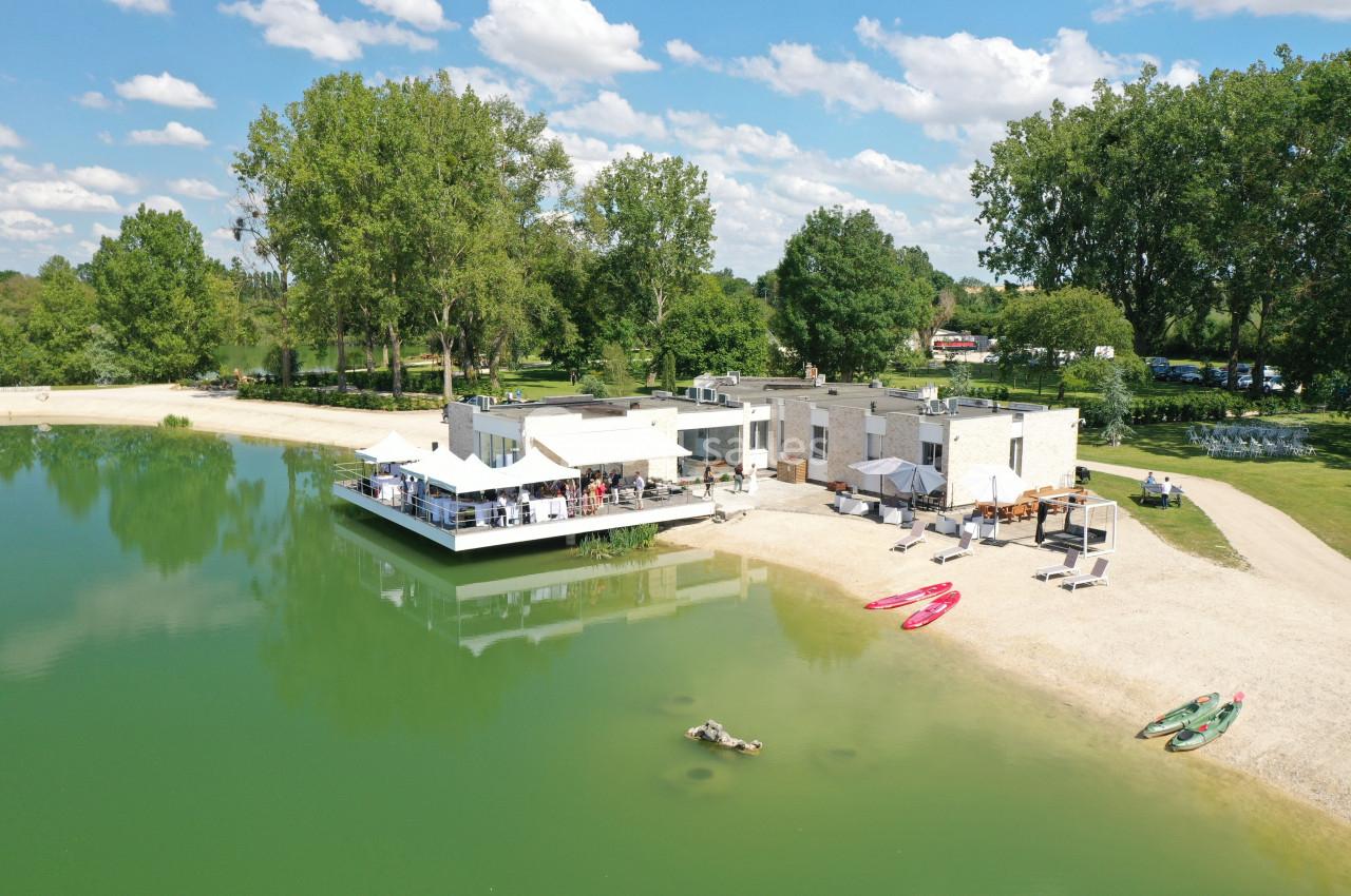 Vue aérienne d'un bâtiment moderne au bord d'un lac avec une plage, des kayaks et des arbres en arrière-plan.