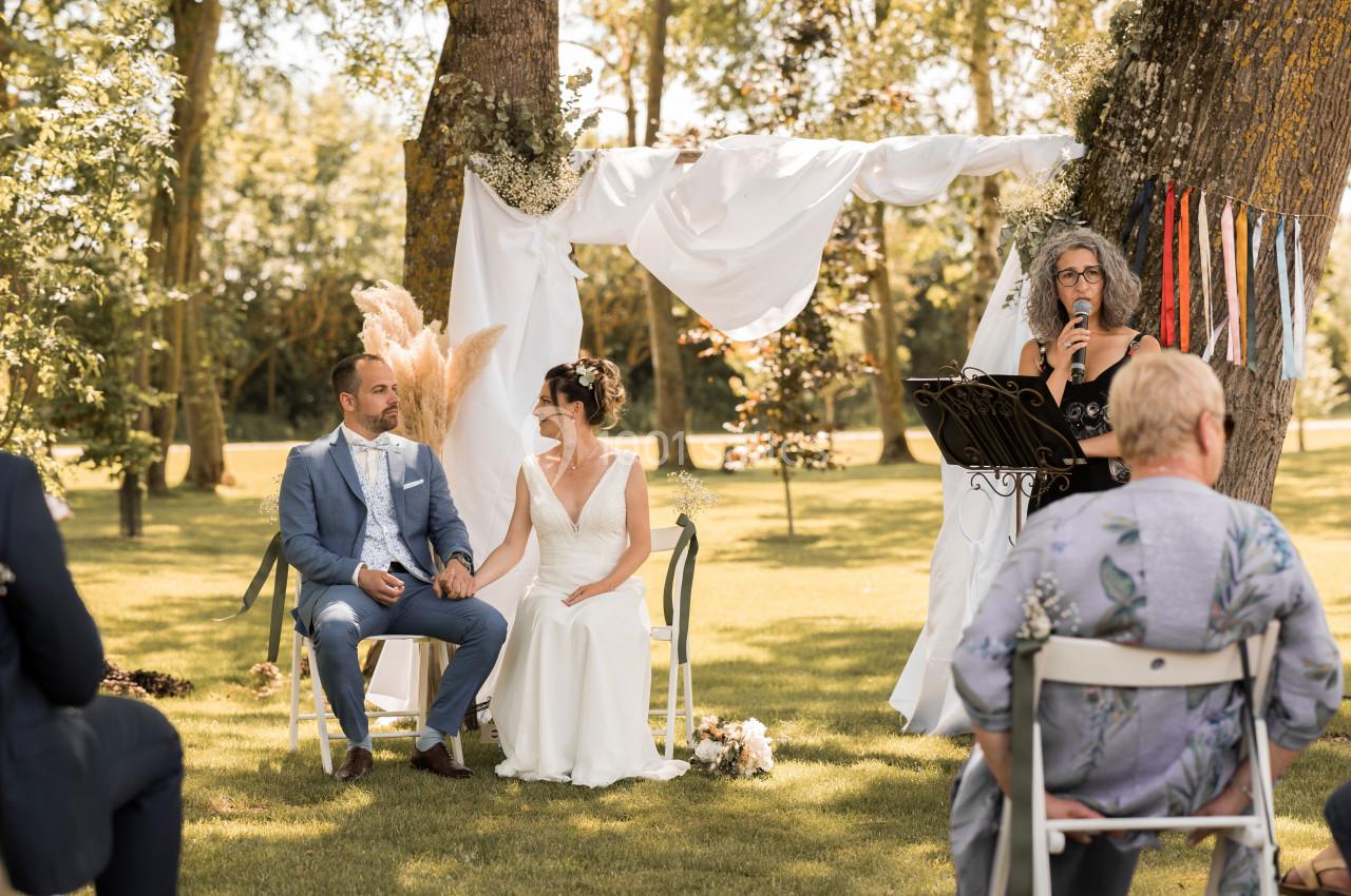 Un couple assis lors d'une cérémonie en plein air, écoutant une femme lire un discours devant une arche décorée.
