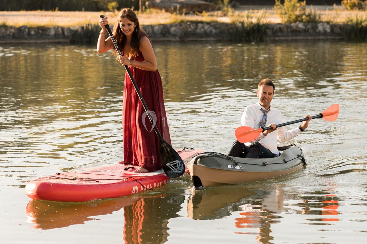 Une femme en robe rouge sur une planche de paddle et un homme en kayak pagaient sur un lac entouré d'arbres.
