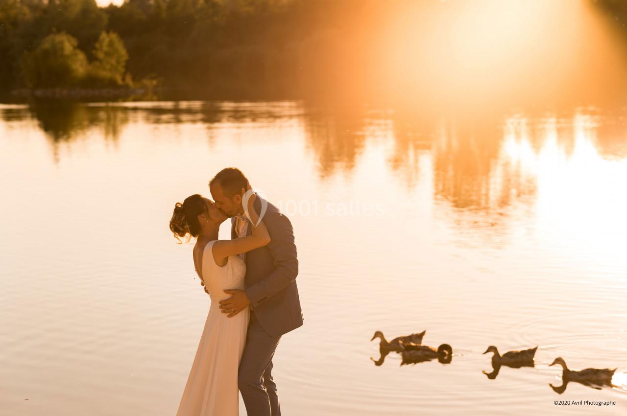 Un couple s'embrasse au bord d'un lac au coucher du soleil, avec des canards nageant sur l'eau calme.
