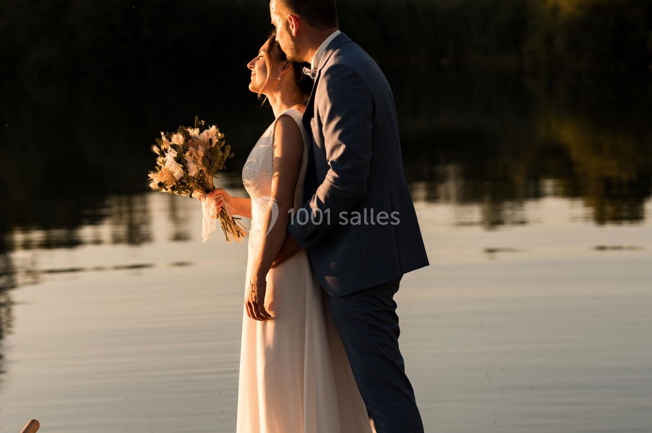 Un couple en tenue de mariage se tient sur un ponton en bois au bord d'un lac, éclairé par une lumière dorée.