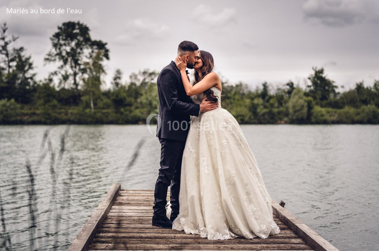 Un couple de mariés s'embrassant sur un ponton en bois au bord d'un lac, sous un ciel nuageux.