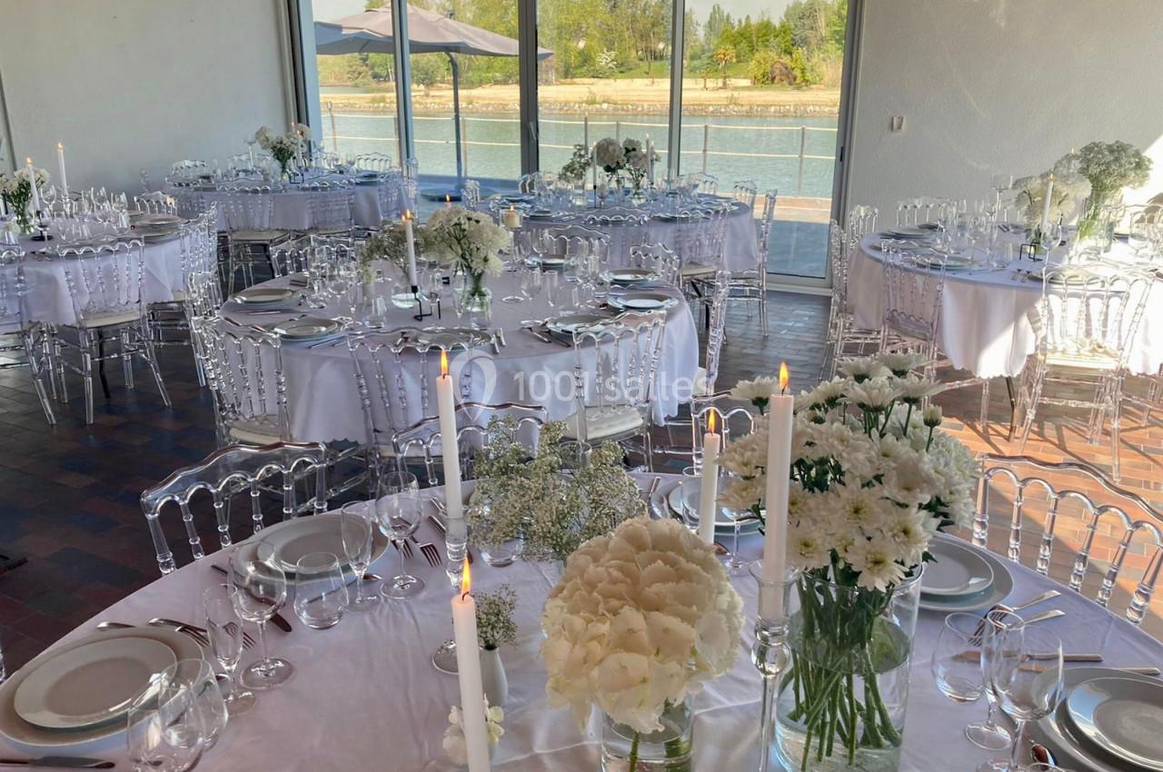 Salle de réception lumineuse avec tables rondes dressées, décorées de fleurs blanches et bougies, vue sur un plan d'eau.