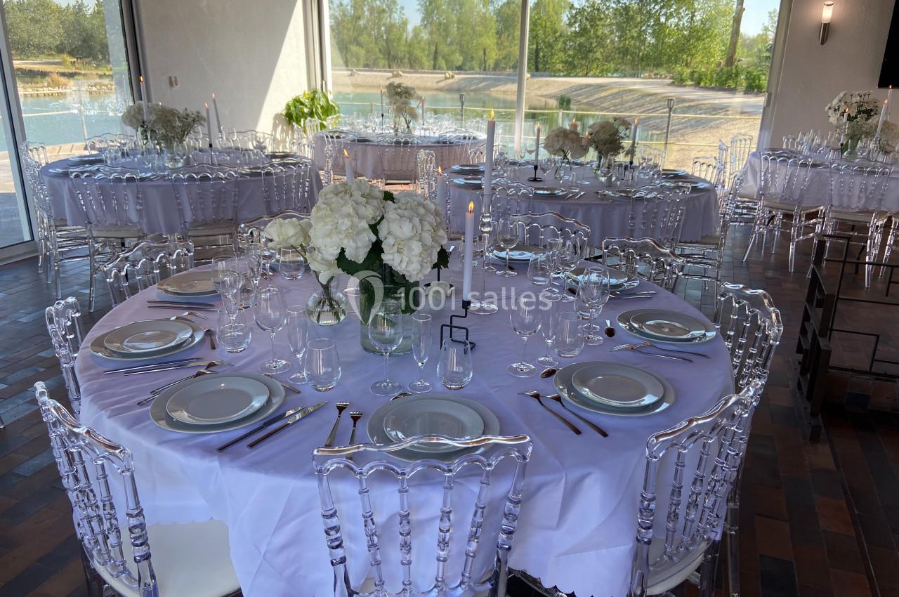 Tables rondes dressées avec nappes blanches, vaisselle élégante et bouquets de fleurs blanches dans une salle lumineuse.