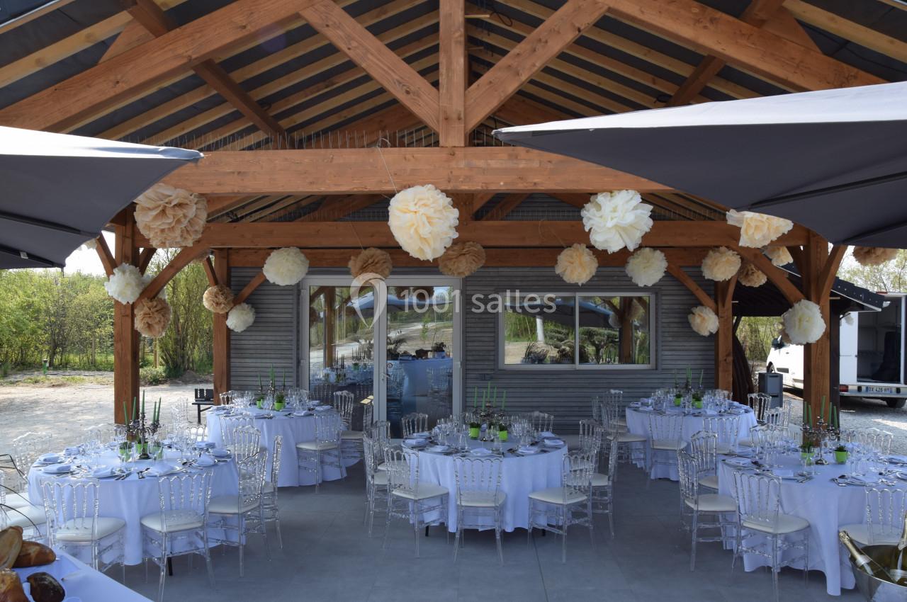 Salle de réception en plein air avec charpente en bois, tables rondes dressées et décorations suspendues.