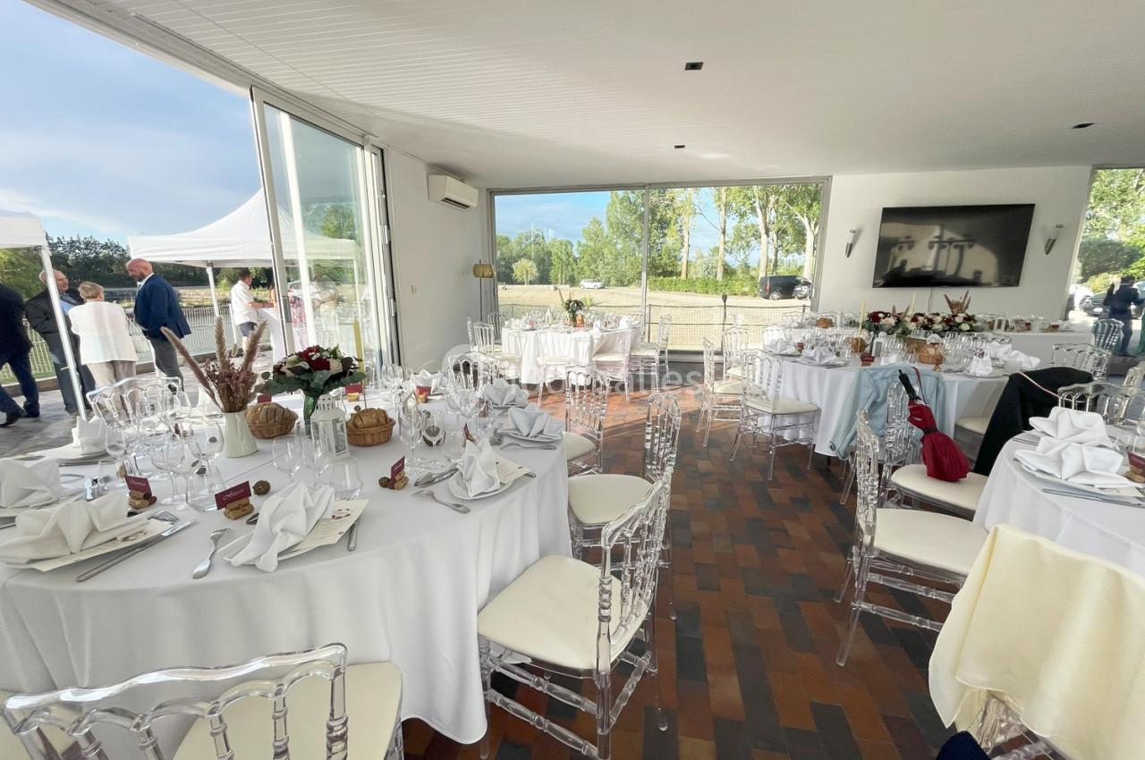 Salle lumineuse avec tables rondes dressées pour un événement, chaises transparentes et vue sur un extérieur verdoyant.