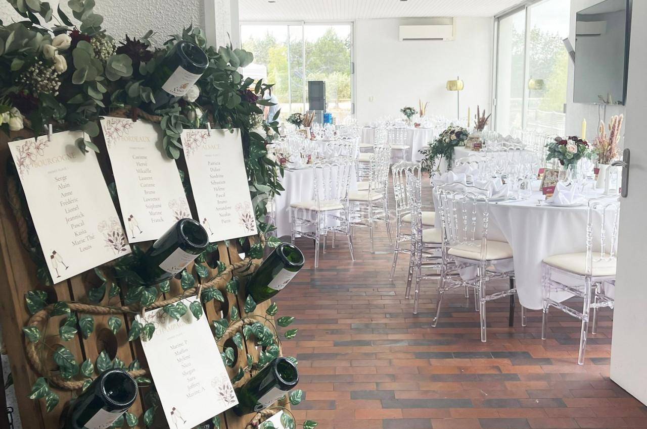 Salle de réception décorée pour un mariage avec tables dressées et plan de table affiché sur un support en bois.