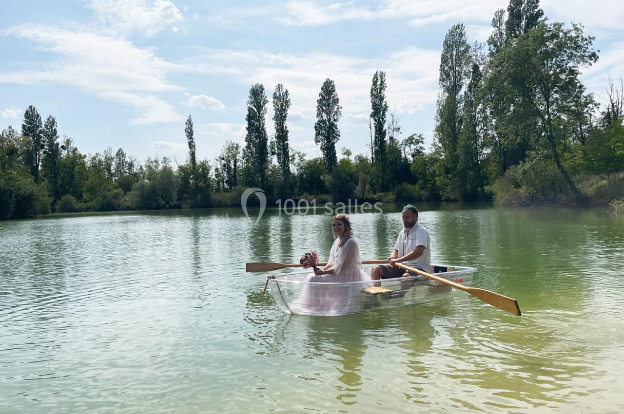 Un couple assis dans une barque transparente rame sur un lac entouré d'arbres par une journée ensoleillée.