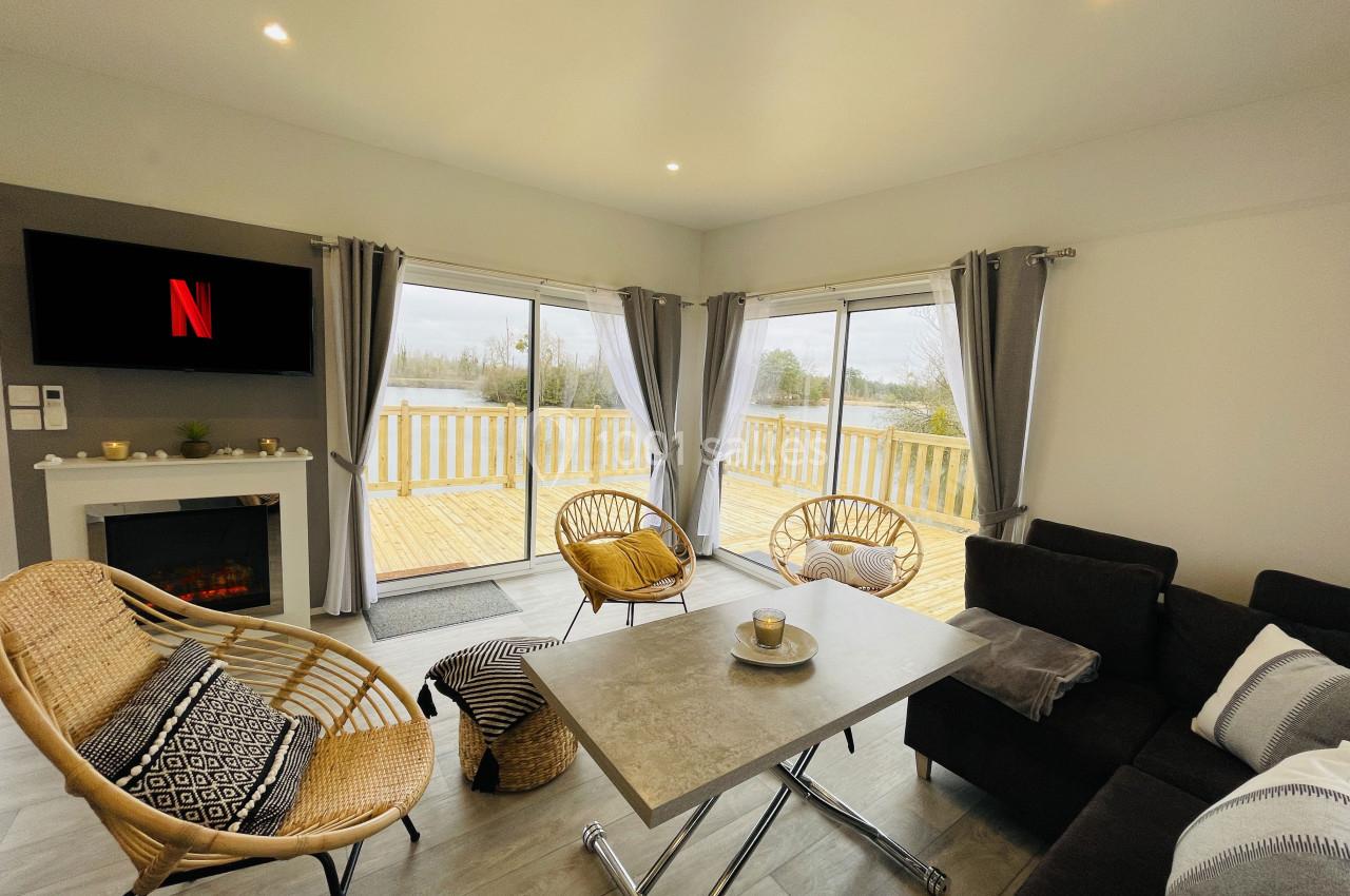 Salon lumineux avec canapé, fauteuils en rotin, table basse, cheminée électrique et grandes baies vitrées donnant sur une…