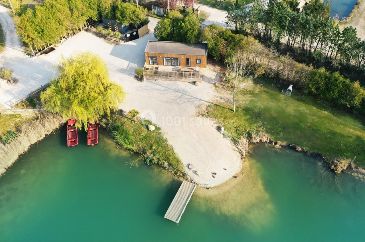 Vue aérienne d'une maison en bois près d'un lac avec un ponton, deux barques rouges et un espace arboré.