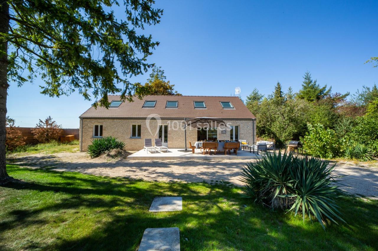 Maison moderne avec toit en pente, terrasse aménagée et jardin arboré sous un ciel bleu clair.