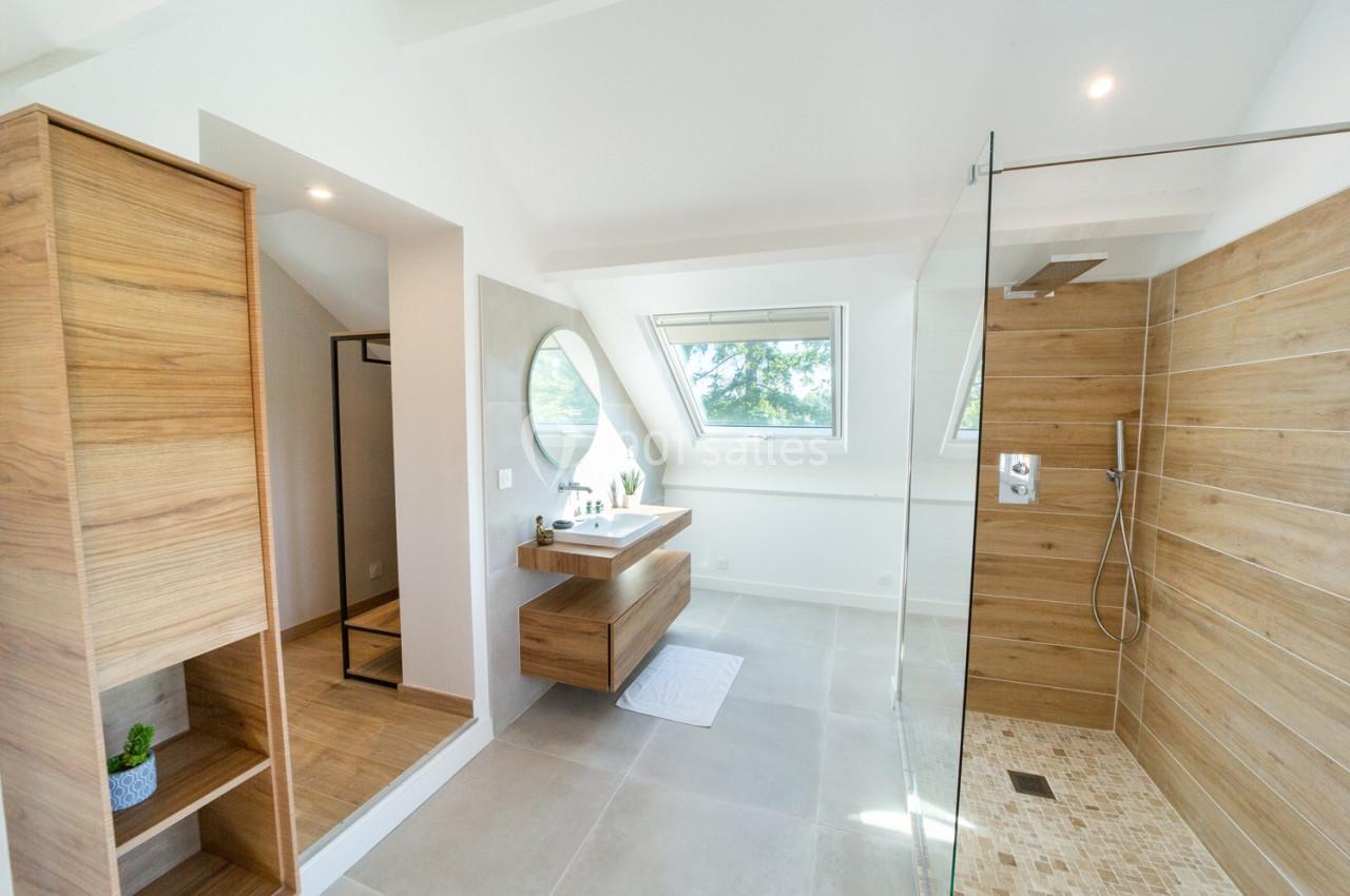 Salle de bain moderne avec douche à l'italienne, meuble en bois clair, miroir rond et lumière naturelle.