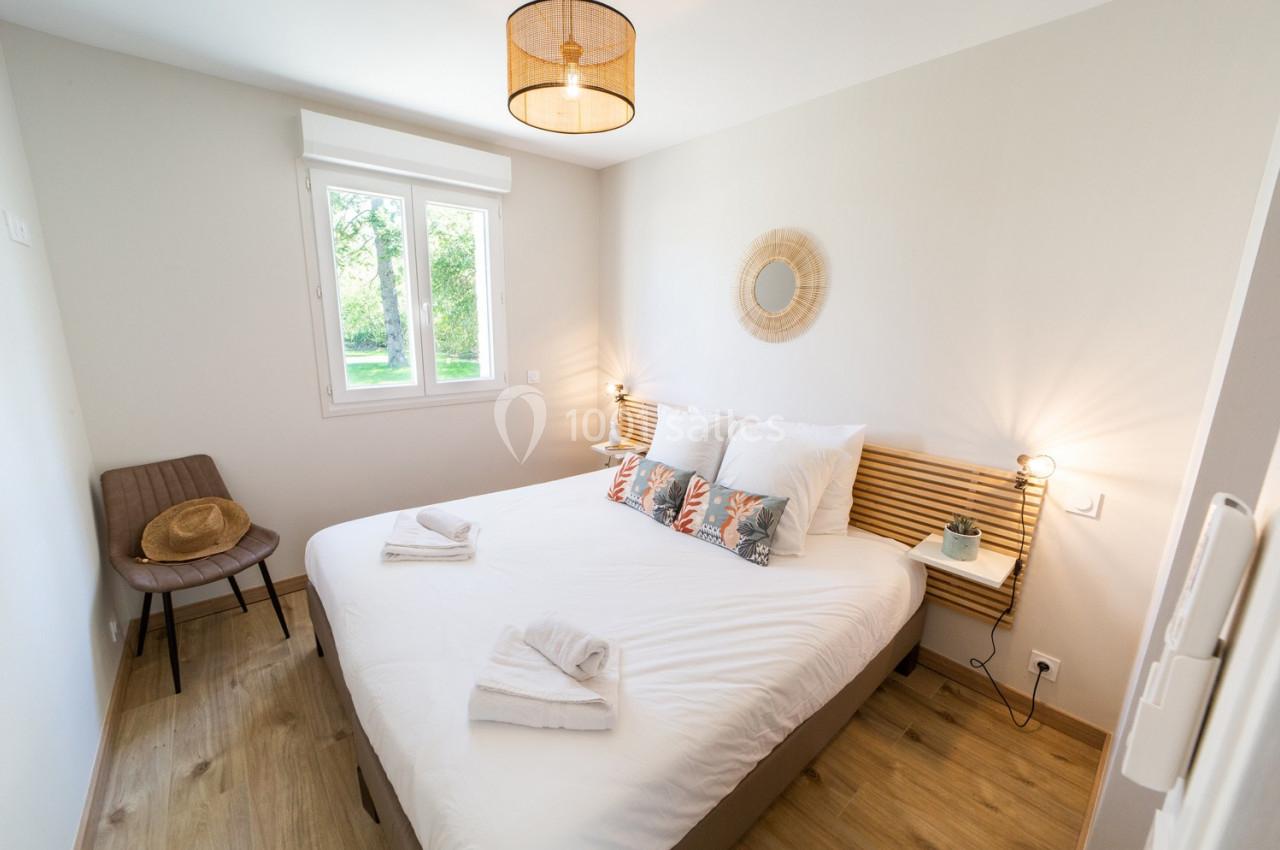 Chambre lumineuse avec lit double, draps blancs, deux lampes de chevet, chaise et fenêtre donnant sur un jardin.