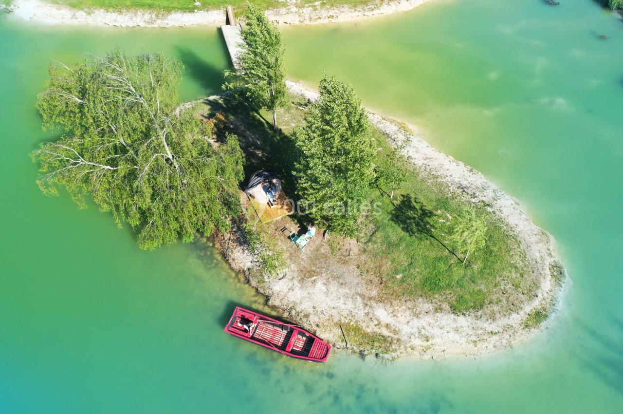 Vue aérienne d'une petite île verdoyante avec un bateau rouge amarré et un ponton en bois sur un lac turquoise.