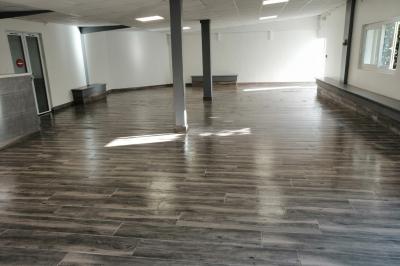 Salle vide avec un sol en parquet sombre, murs blancs, fenêtres et piliers gris sous un plafond à dalles blanches.