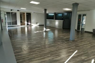 Salle vide avec un sol en parquet sombre, murs blancs, fenêtres et piliers gris sous un plafond à dalles blanches.