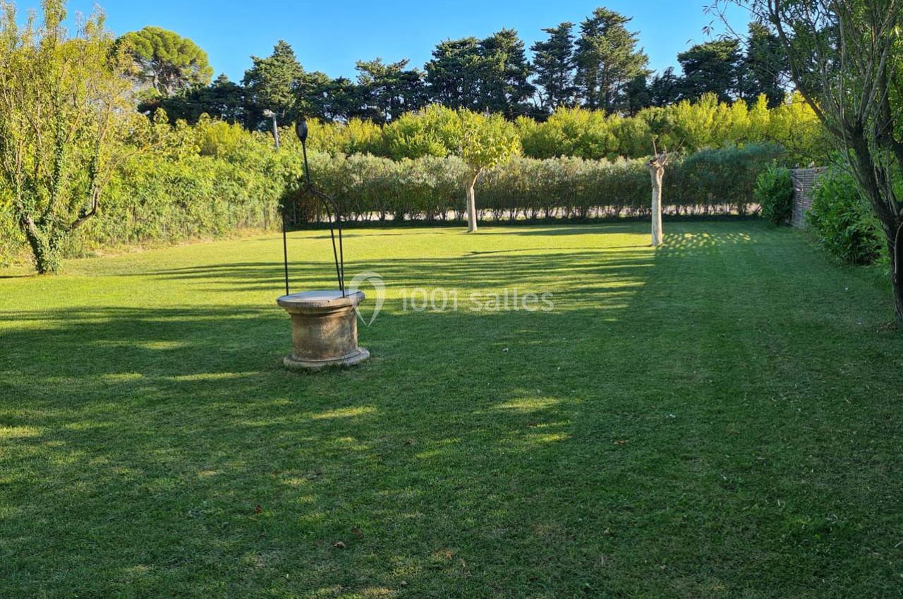 Puits en pierre au centre d'une pelouse verdoyante entourée d'arbres et de haies sous un ciel dégagé.