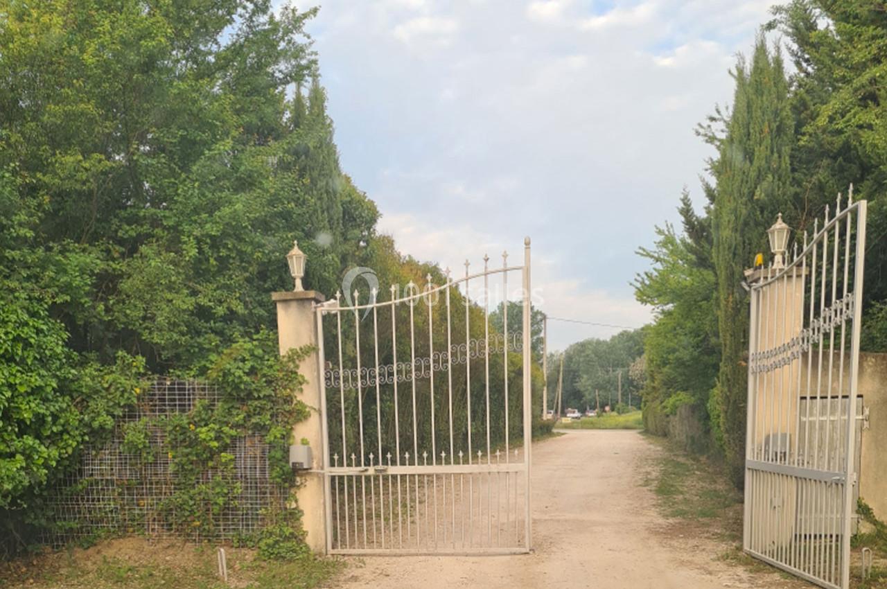 Portail métallique blanc ouvert donnant sur un chemin de terre bordé de végétation et d'arbres.