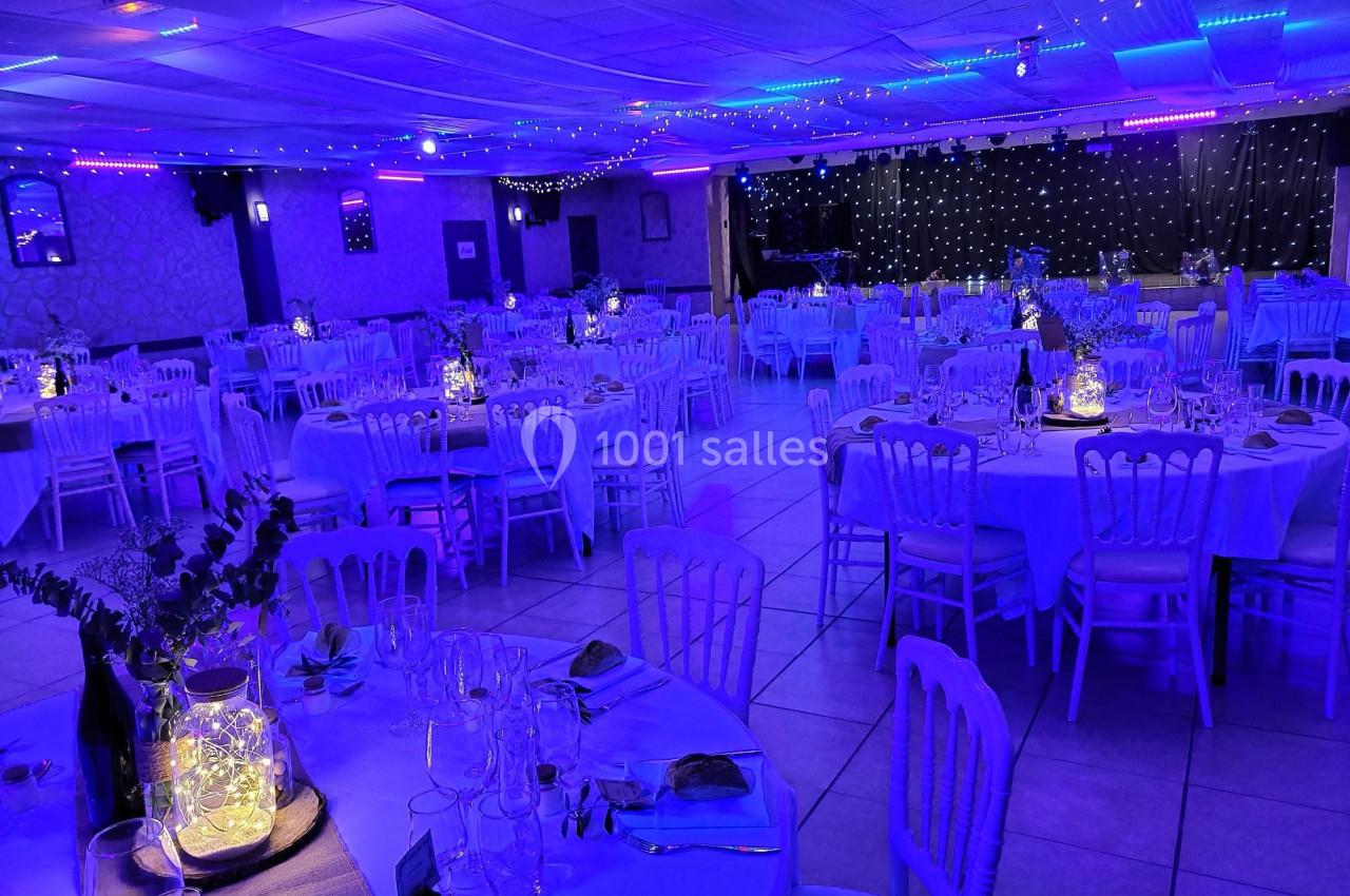 Salle de réception décorée avec des tables rondes dressées, éclairage bleu et guirlandes lumineuses au plafond.