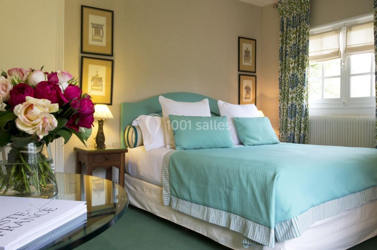 Chambre lumineuse avec lit double, couvre-lit turquoise, fleurs sur une table et fenêtre encadrée de rideaux à motifs.