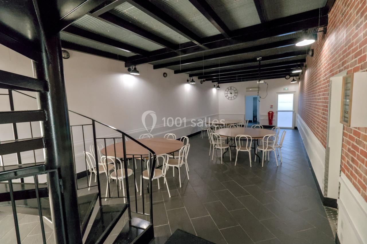 Salle avec des tables rondes et des chaises blanches, éclairée par des lampes suspendues, avec un escalier en colimaçon au…